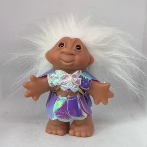 May include: A vintage troll doll with a tan face, brown eyes, and white, spiky hair. The doll wears a purple and iridescent outfit with a bow and gold accents. The doll is standing with its arms outstretched.