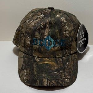 May include: Camouflage baseball cap with the word "DODGE" embroidered in teal. The cap features a Realtree Xtra tag, and is made of a camouflage pattern in shades of brown, green, and tan.