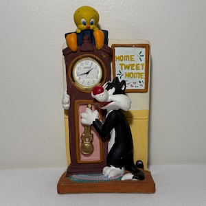 Puede incluir: Un reloj decorativo de mesa con diseño de reloj de pie marrón y esfera dorada. El gato Silvestre está de pie a la derecha, mientras que Piolín se sienta en la parte superior. Un letrero en la pared dice HOME TWEET HOME en letras amarillas.