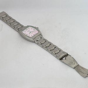 Pode incluir: Um rel&oacute;gio de pulso prateado com um mostrador quadrado e uma pulseira de metal prateado. O mostrador do rel&oacute;gio tem um fundo branco com n&uacute;meros e ponteiros pretos.