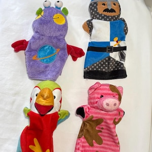 Op de afbeelding: Vier kleurrijke handpoppen: een paarse alien met een planeetontwerp, een ridder in blauw en wit, een papegaai in rood en groen en een roze varken met bruine moddervlekken. Deze pluche poppen zijn ontworpen voor kinderspel.