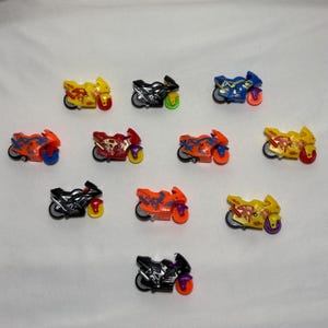 May include: A selection of toy motorbikes in diverse colours, such as yellow, black, red, blue, and orange. Each miniature bike has detailed designs and contrasting wheel colours, arranged on a white surface.
