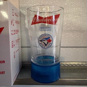 May include: Clear Budweiser pint glass with a blue base. The glass features the Budweiser logo in red and white, along with the Toronto Blue Jays logo. The text "Proud Partner Fier Partenaire" is also visible.