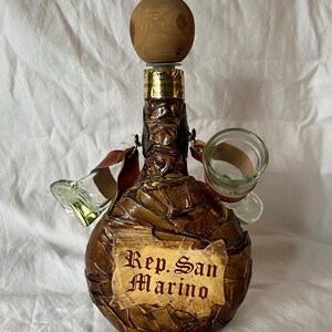 Puede incluir: Una botella de cuero marrón envuelta en cuero con un tapón de madera y dos pequeños vasos de chupito de vidrio unidos. La botella tiene una etiqueta que dice "Rep. San Marino".