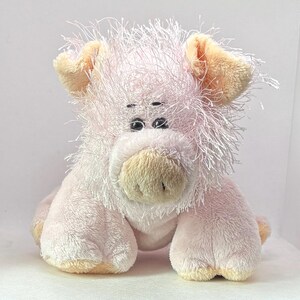 May include: A plush pig toy in shades of pink and cream. The pig has a shaggy, textured mane and ears, with small black eyes and a cream-colored snout. The toy is sitting upright, with its front paws visible.