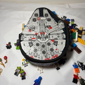 Op de afbeelding: Een zwarte en grijze lunchtas met een gedetailleerde afbeelding van de Millennium Falcon uit Star Wars. De tas heeft een zwarte rits en een riem. Verschillende Lego-minifiguren en Lego-stenen liggen rond de tas.