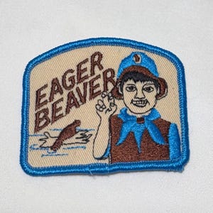 May include: A rectangular embroidered patch with a beige background and a blue border. It features a brown beaver swimming in blue water and a person wearing a blue hat and scarf with the text EAGER BEAVER embroidered in brown letters.