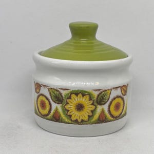 May include: A white ceramic sugar bowl with a green lid. The bowl features a floral design with yellow sunflowers, green leaves, and brown accents. The lid has a rounded knob and a ridged texture.