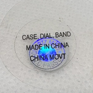 Pode incluir: Close-up de um adesivo redondo com o texto "CASE, DIAL, BAND" e "MADE IN CHINA" em letras pretas. O adesivo tamb&eacute;m tem um c&iacute;rculo azul com o texto "CHINA MOVT" em letras brancas.