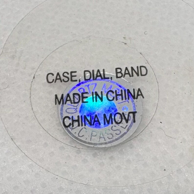 Pode incluir: Close-up de um adesivo redondo com o texto "CASE, DIAL, BAND" e "MADE IN CHINA" em letras pretas. O adesivo tamb&eacute;m tem um c&iacute;rculo azul com o texto "CHINA MOVT" em letras brancas.