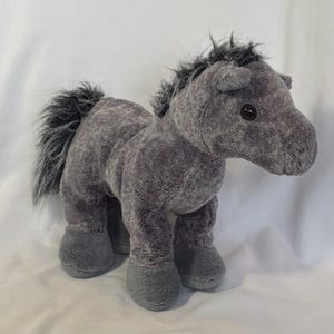 Op de afbeelding: Een grijs pluche paardenspeelgoed met een lange, vloeiende manen en staart. Het paard heeft een vriendelijk gezicht met zwarte ogen en een zwarte neus.
