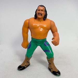May include: A vintage wrestling action figure with orange skin, green trousers with purple detailing, and brown boots. The figure has black hair and a moustache. The toy is standing on a white surface.