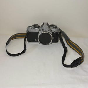 May include: A silver and black Nikon 35mm film camera body with a circular lens cap attached to the front. The camera features a top-mounted silver prism housing with the Nikon logo and is connected to a grey and black neck strap with yellow trim.