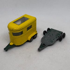 May include: A vintage yellow toy horse trailer with black wheels and a green hitch. Beside it is a green and gray flatbed trailer with black wheels. The toy trailers are on a light background.