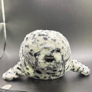 Puede incluir: Un peluche de foca con un patrón de manchas blancas y negras. El juguete tiene una nariz negra, bigotes y aletas pequeñas. La foca está tumbada sobre una superficie oscura.