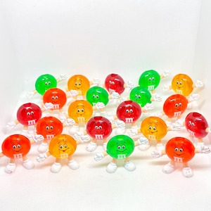 May include: A collection of colorful, anthropomorphic candy figurines. Each figurine has a round, glossy candy body in various colors like red, green, orange, and yellow. They have white arms, legs, and feet, and a smiling face with the letter "M".