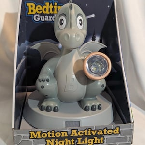 May include: A gray dragon-shaped night light with a round, tan-colored light. The night light is motion-activated and sits on a gray base. The box reads "Bedtime Guard" and "Motion Activated Night Light".