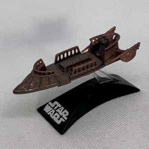 May include: A brown Star Wars model of a vehicle, with a detailed design. The model is mounted on a black base with the Star Wars logo in white. The vehicle has a cockpit and side panels.