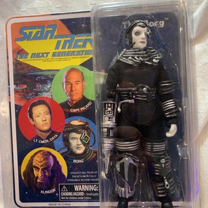 Peut inclure: Une figurine d'action Star Trek: La Nouvelle Génération d'un Borg portant une combinaison noire et argentée, un casque noir avec une visière argentée et une arme argentée et noire. La figurine est emballée dans une boîte blister en plastique transparent avec un dos en carton. Le dos présente des images des personnages de la série, notamment le Capitaine Picard, le Lieutenant-Commandant Data et Klingon. Le texte au dos indique "Star Trek: La Nouvelle Génération" et "Borg".