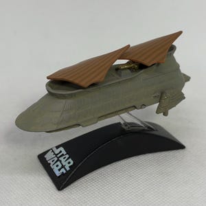May include: A Star Wars model of a vehicle in shades of green and brown, with two brown wing-like structures. The model is mounted on a black base with the Star Wars logo in white.