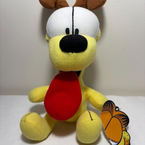 May include: A yellow plush dog toy with a red tongue, brown ears, and a black nose. The dog has a white head and a tag with a cartoon image. The toy is soft and cuddly.