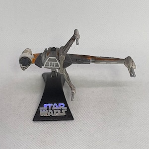 May include: A miniature Star Wars B-wing starfighter model. The model is primarily gray with orange and brown accents. It is mounted on a black stand with the Star Wars logo in silver.