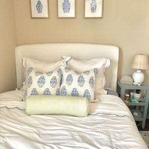 Op de afbeelding: Een wit bed met een wit hoofdbord en witte beddengoed. Er zijn twee blauwe en groene gepatroneerde kussens en een groen gestreept kussen op het bed. Drie ingelijste prints van blauwe en witte gembervazen hangen aan de muur boven het bed.