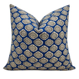 May include: A square throw pillow featuring a vibrant blue background with a repeating pattern of cream-colored, petal-shaped motifs. Each motif contains a central eye-like design outlined in black, creating a consistent, symmetrical graphic print.