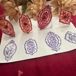 May include: A collection of floral design stamps in various shapes and sizes. The stamps are displayed on clear acrylic stands, with impressions in purple and brown ink on white paper. The designs include oval and rectangular frames with floral motifs.