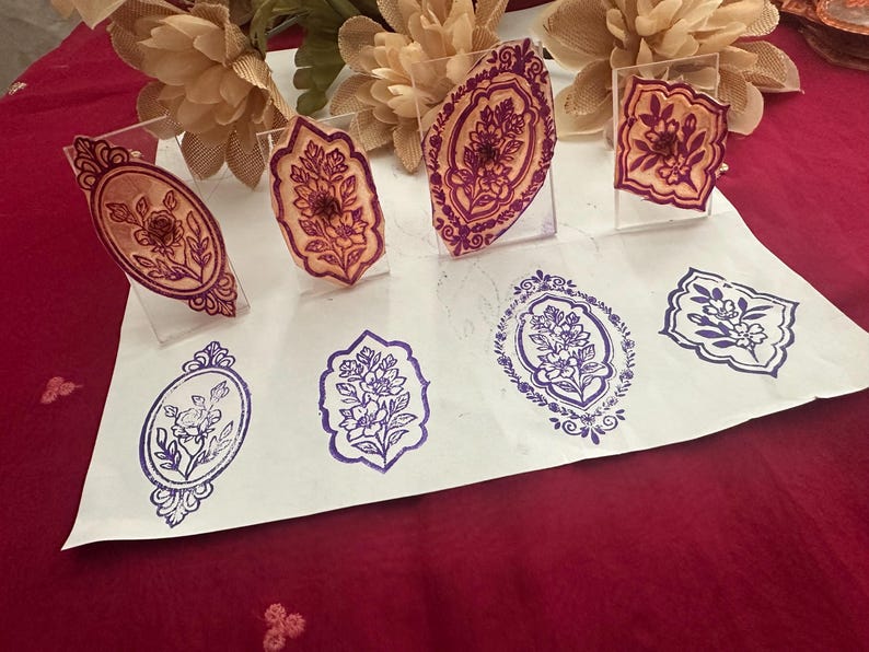 May include: A collection of floral design stamps in various shapes and sizes. The stamps are displayed on clear acrylic stands, with impressions in purple and brown ink on white paper. The designs include oval and rectangular frames with floral motifs.