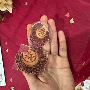 May include: Two rubber stamps with a crescent moon and star design, held against a red background. The stamps are brown with intricate floral and geometric patterns. A sheet of paper with blue stamped designs is visible.