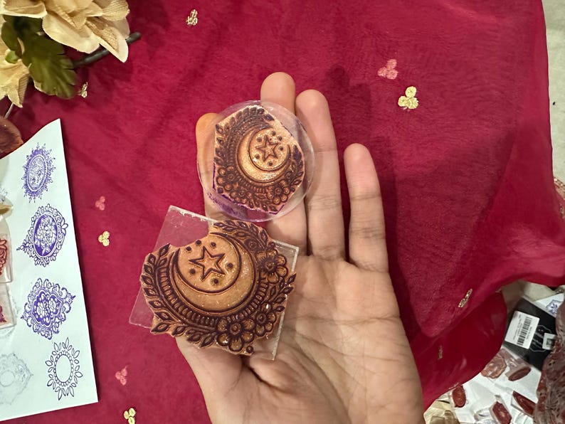 May include: Two rubber stamps with a crescent moon and star design, held against a red background. The stamps are brown with intricate floral and geometric patterns. A sheet of paper with blue stamped designs is visible.