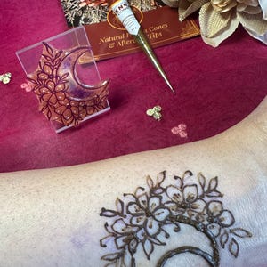 May include: A henna tattoo design featuring a crescent moon and floral elements is visible on the skin. A clear acrylic stamp with a similar design sits next to a henna cone and a brochure. The background is a deep pink.