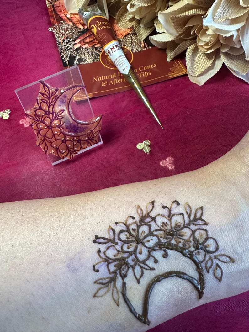 May include: A henna tattoo design featuring a crescent moon and floral elements is visible on the skin. A clear acrylic stamp with a similar design sits next to a henna cone and a brochure. The background is a deep pink.