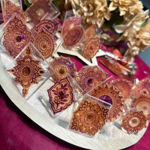 May include: A display of decorative resin pieces with intricate henna-style designs in shades of gold and purple. The pieces are arranged on a wooden tray, with a backdrop of cream-colored flowers and a pink surface.