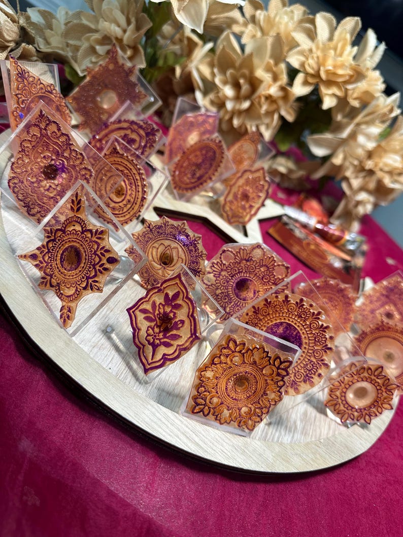 May include: A display of decorative resin pieces with intricate henna-style designs in shades of gold and purple. The pieces are arranged on a wooden tray, with a backdrop of cream-colored flowers and a pink surface.