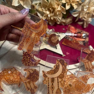May include: Clear resin ornaments with intricate henna-style designs in shades of orange and brown. The ornaments feature crescent moons, floral patterns, and hanging lanterns. Some are held by a hand, while others are displayed on a wooden surface.