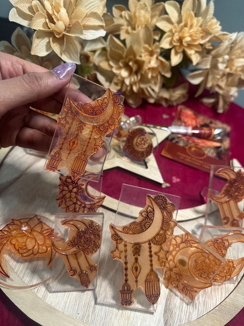 May include: Clear resin ornaments with intricate henna-style designs in shades of orange and brown. The ornaments feature crescent moons, floral patterns, and hanging lanterns. Some are held by a hand, while others are displayed on a wooden surface.