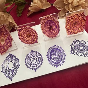 May include: A collection of decorative stamps with intricate floral and geometric designs. The stamps are mounted on clear acrylic blocks, with impressions in shades of purple and orange. The stamped images are displayed on a white surface.