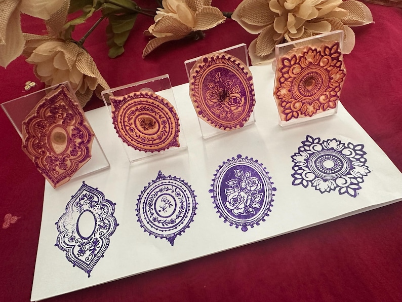 May include: A collection of decorative stamps with intricate floral and geometric designs. The stamps are mounted on clear acrylic blocks, with impressions in shades of purple and orange. The stamped images are displayed on a white surface.