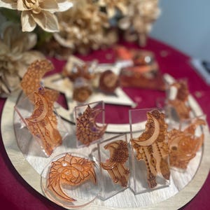 May include: A collection of orange resin art pieces, including crescent moons, floral designs, and other decorative shapes, displayed on a wooden tray. The pieces are set against a deep red background, with blurred floral accents.
