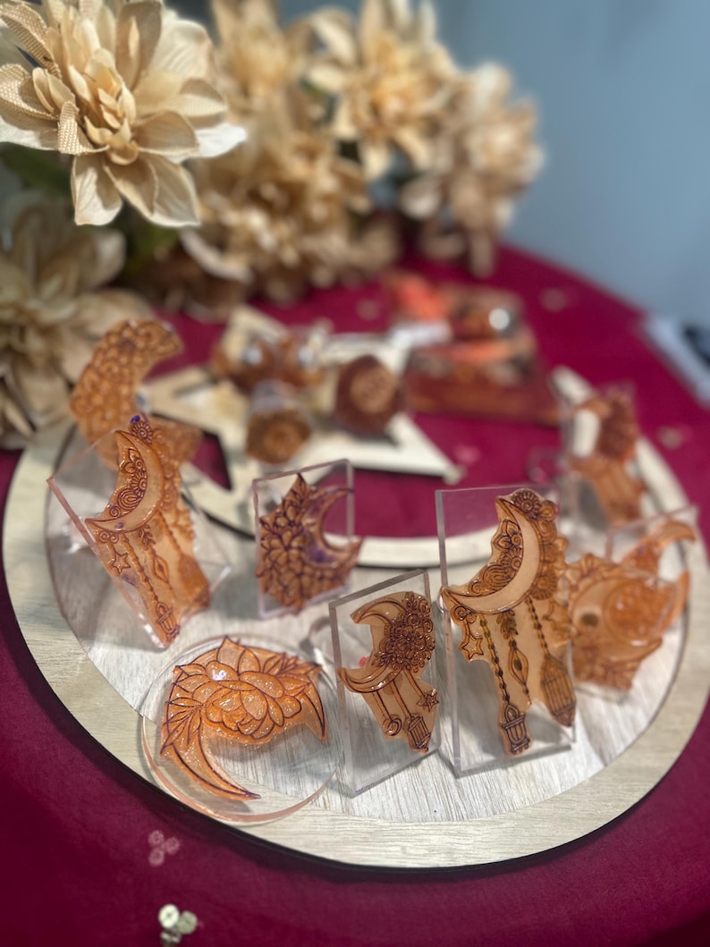 May include: A collection of orange resin art pieces, including crescent moons, floral designs, and other decorative shapes, displayed on a wooden tray. The pieces are set against a deep red background, with blurred floral accents.