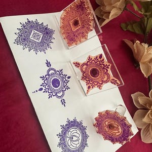 May include: A white sheet of paper with purple mandala designs stamped on it. Clear acrylic stamps with similar designs are next to the stamped images. The background is a deep red color with a few decorative flowers.