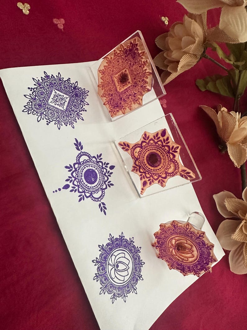 May include: A white sheet of paper with purple mandala designs stamped on it. Clear acrylic stamps with similar designs are next to the stamped images. The background is a deep red color with a few decorative flowers.
