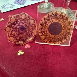 May include: Two decorative stamps with intricate floral mandala designs. The stamps are made of a clear material with a brown ink design. The stamps are on a red surface. The image also shows stamped impressions in blue ink on a white surface.