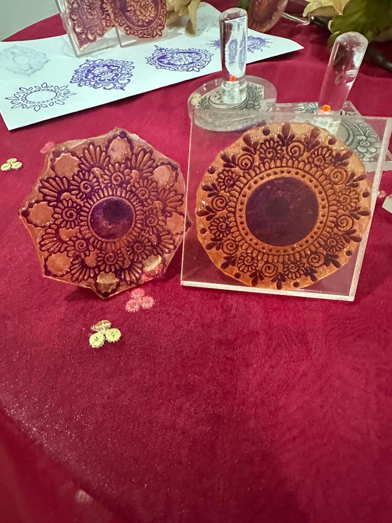 May include: Two decorative stamps with intricate floral mandala designs. The stamps are made of a clear material with a brown ink design. The stamps are on a red surface. The image also shows stamped impressions in blue ink on a white surface.