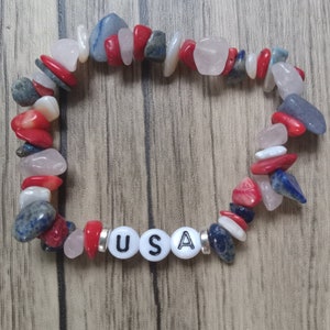 May include: A beaded bracelet featuring red, white, and blue stones, with the letters "USA" spelled out in white beads. The bracelet is made of various shapes and sizes of stones.