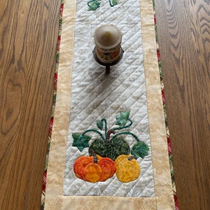 Może przedstawiać: Bieżnik stołowy pikowany z kremowym tłem i wzorem brązowych, zielonych i pomarańczowych dyni. Bieżnik ma beżową obwódkę z czerwoną i zieloną kratą.