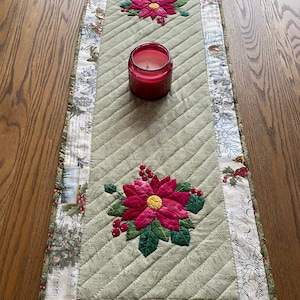 Pode incluir: Um corredor de mesa acolchoado verde com uma borda de estampa floral branca e verde. O corredor de mesa apresenta duas flores de poinsettia bordadas com pétalas vermelhas e folhas verdes. Uma vela vermelha é colocada sobre o corredor de mesa.