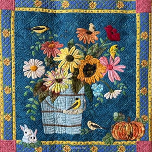 May include: A quilted wall hanging featuring a vibrant floral arrangement in a blue bucket, with sunflowers, daisies, and other colorful flowers. Goldfinches and a pumpkin are also depicted. The quilt is framed with a yellow and blue border.